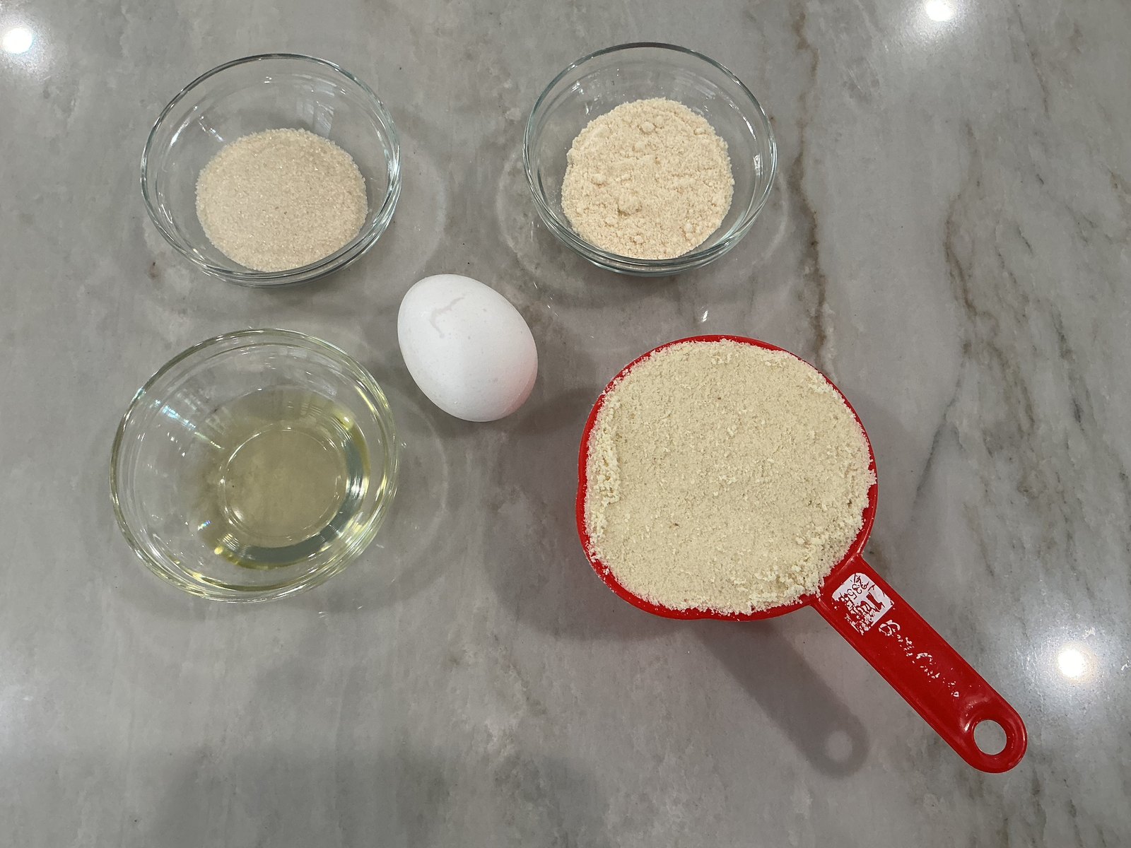 Measured almond flour, coconut flour, sugar, oil, and an egg arranged on a kitchen counter