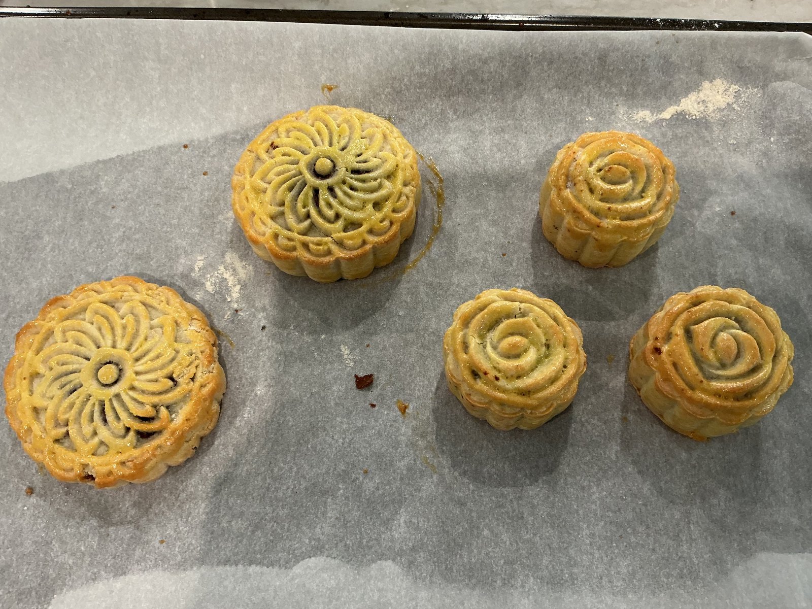 Five golden mooncakes with flower and rose patterns on a parchment-lined baking tray